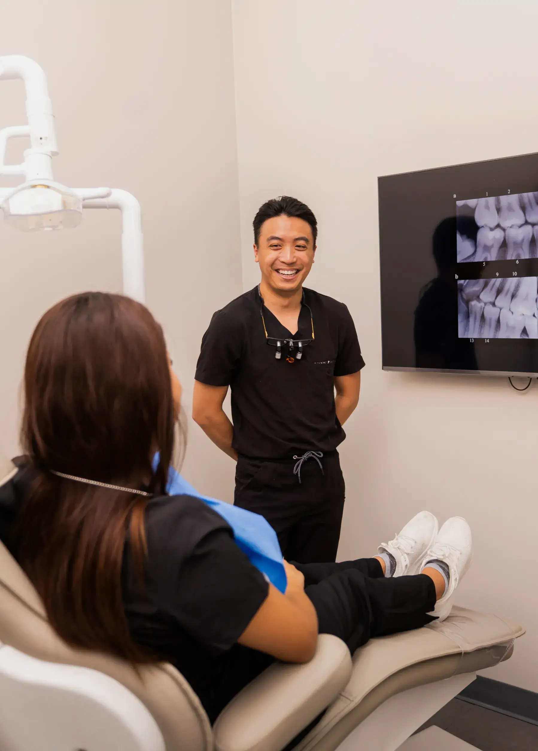 Dental professional smiling at a patient seated in a dental chair, with dental X-ray images on a screen in the background.