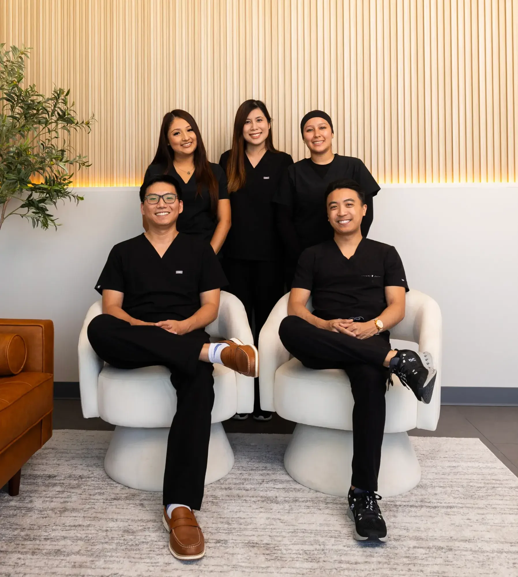 Group of five healthcare professionals in black scrubs posing in a reception area with a wood-panelled wall and a plant nearby.