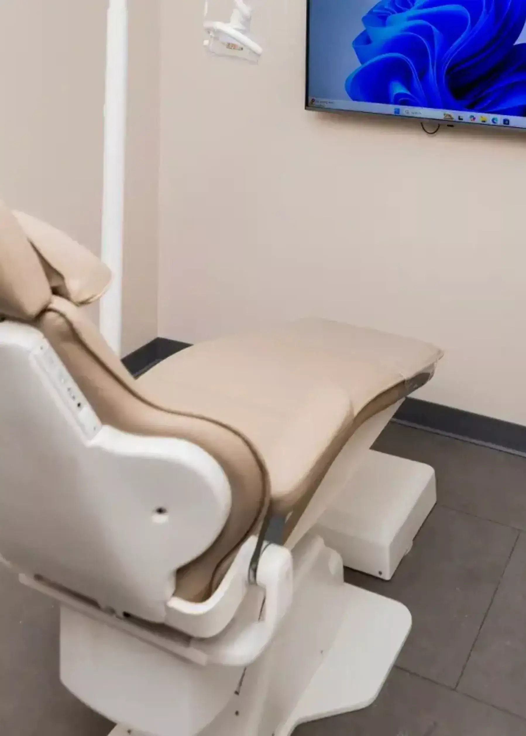 Beige recliner medical chair in a clinic, with a padded headrest and footrest next to a wall-mounted monitor.