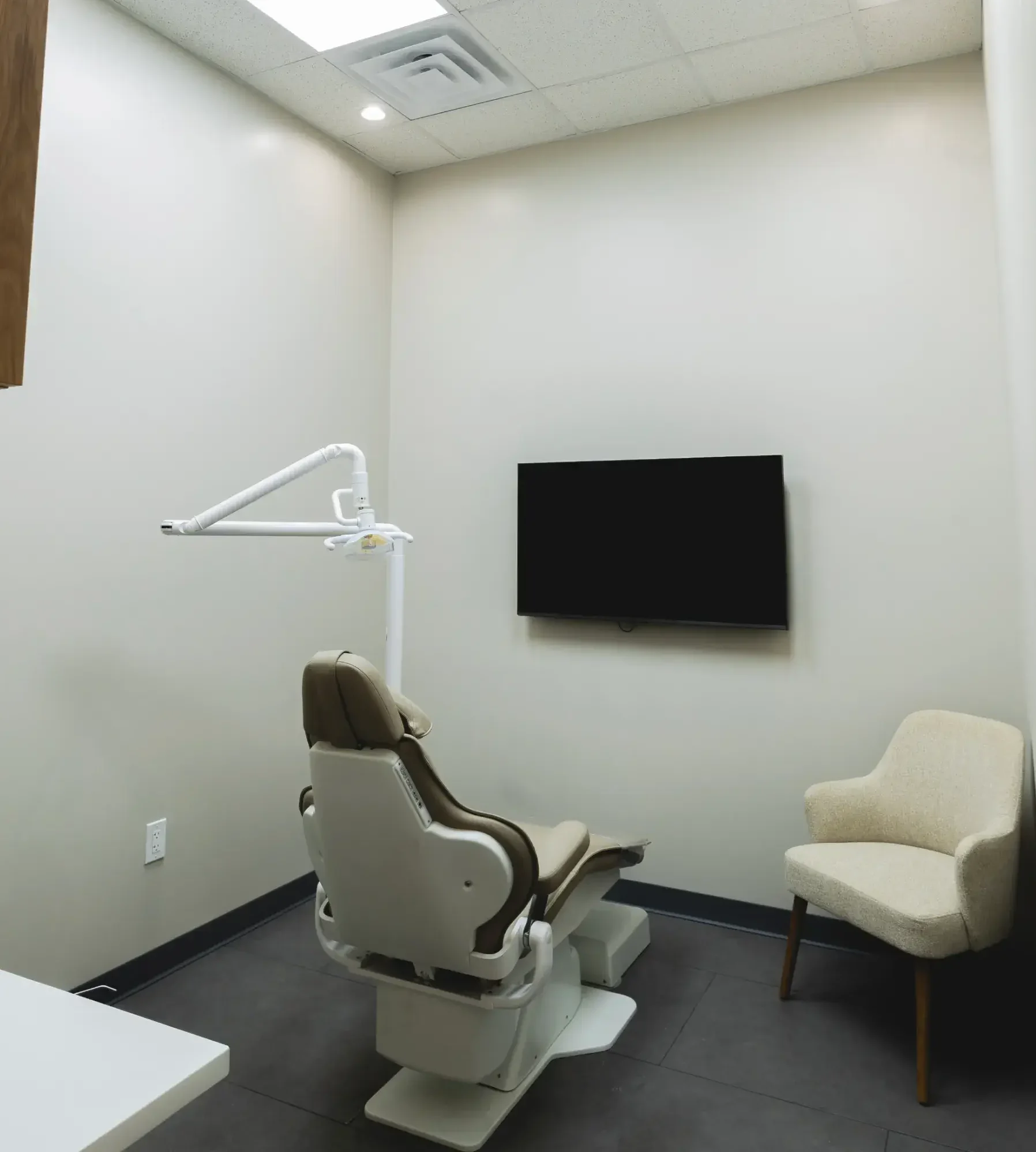 Dental treatment chair in a small clinic room with a wall-mounted monitor and dental equipment nearby.
